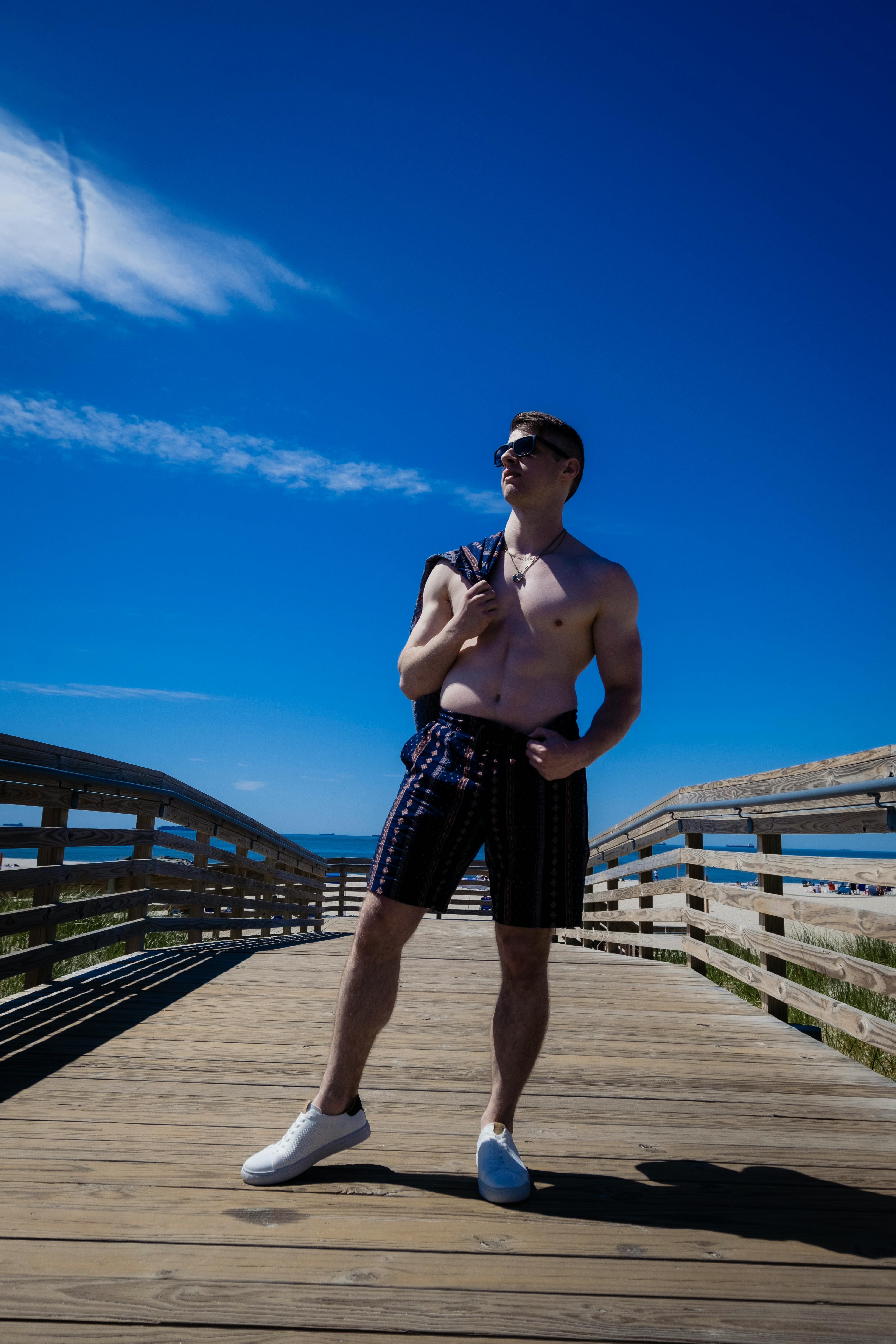 a shirtless man in printed shorts standing on a wooden bridge