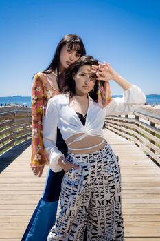 Two stylish women pose on a scenic beach pier under a clear blue sky.