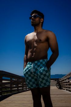Shirtless man wearing sunglasses and checkered shorts stands confidently on a beach boardwalk.