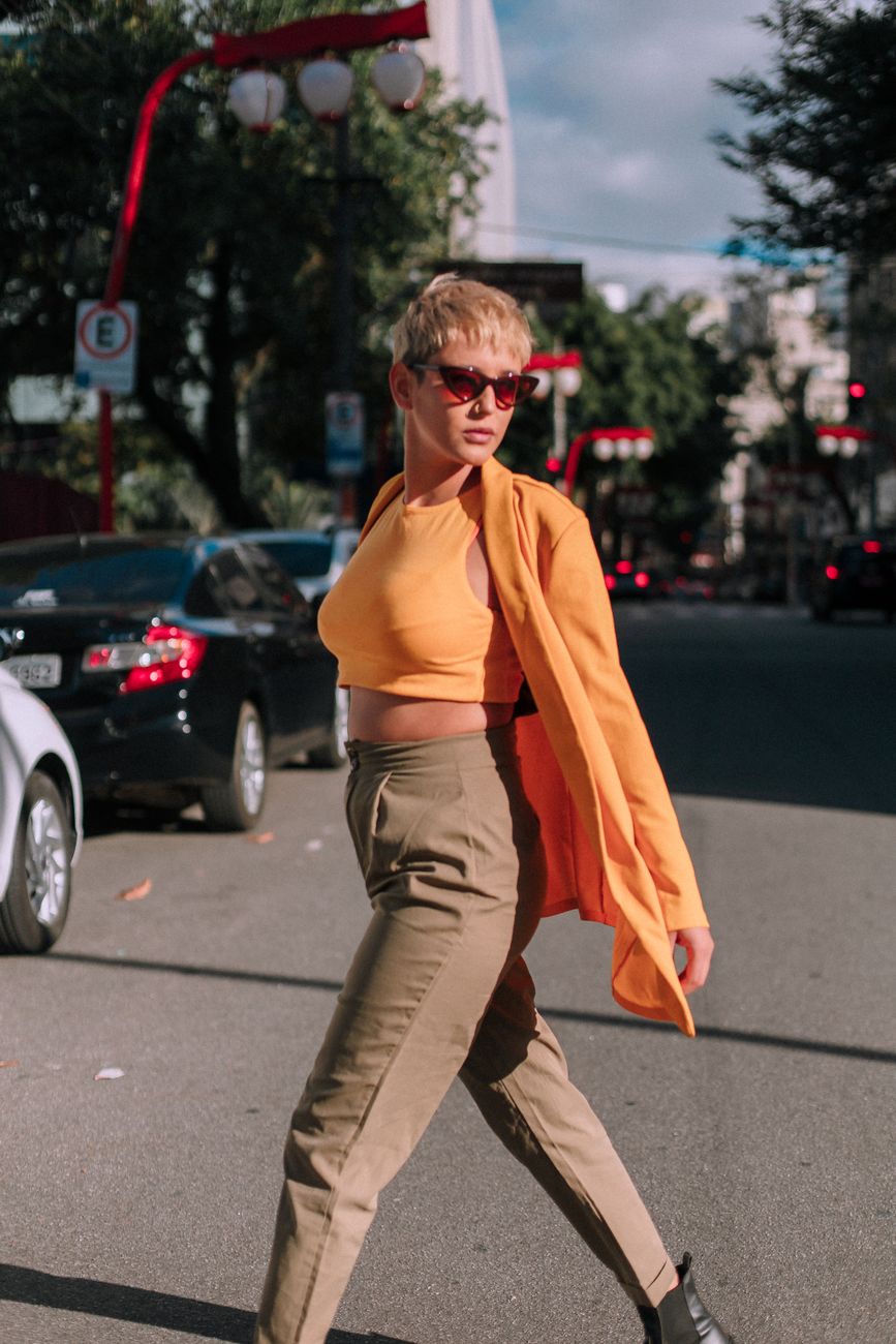 Confident woman in orange crop top and jacket crossing street with sunglasses, showcasing urban fashion.