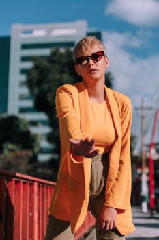 Trendy woman in an orange blazer posing confidently outdoors with sunglasses on a bright day.