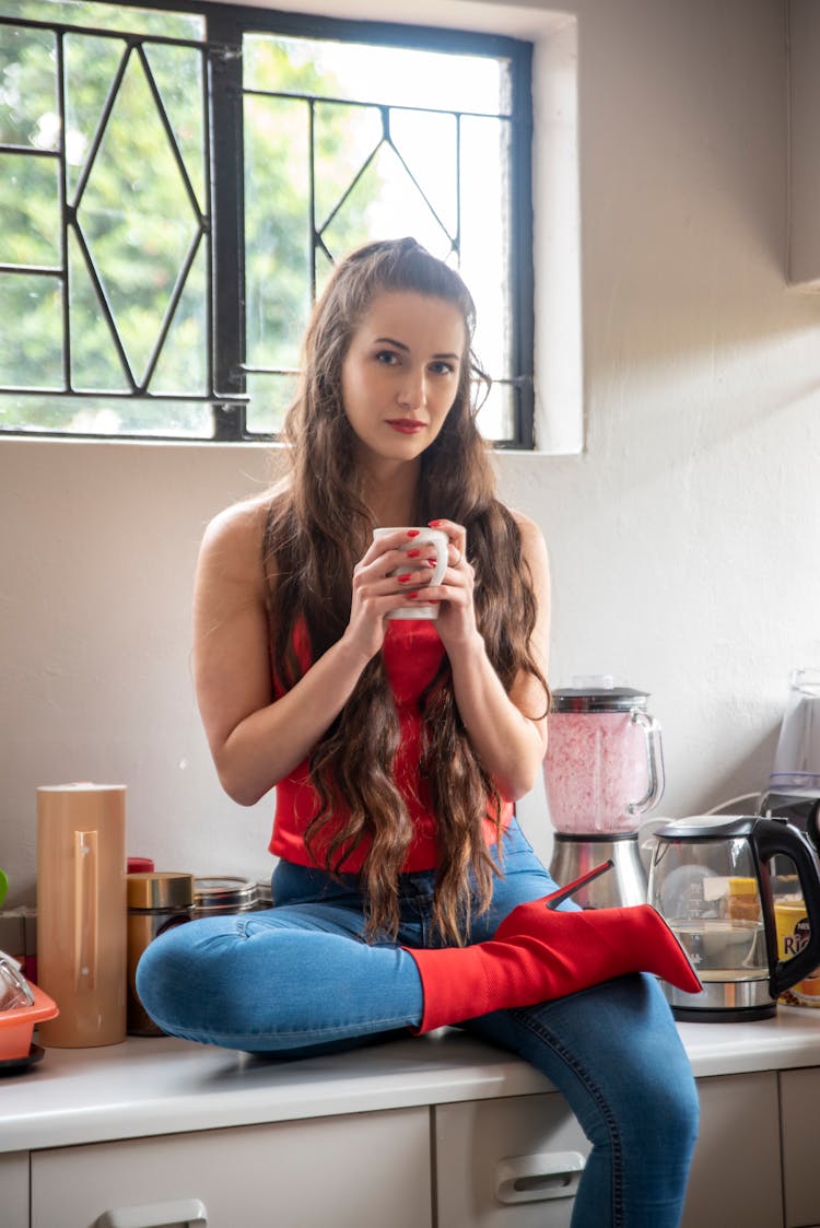 Woman In Red Tube And Blue Denim Jeans Sitting On Kitchen Counter While Holding A Mug 