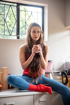 A young woman with long hair sits on a kitchen counter, holding a mug, surrounded by kitchen items.