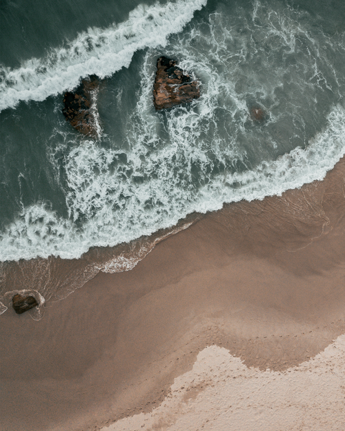 Free A breathtaking aerial view of ocean waves crashing onto a sandy beach with visible rocks. Stock Photo