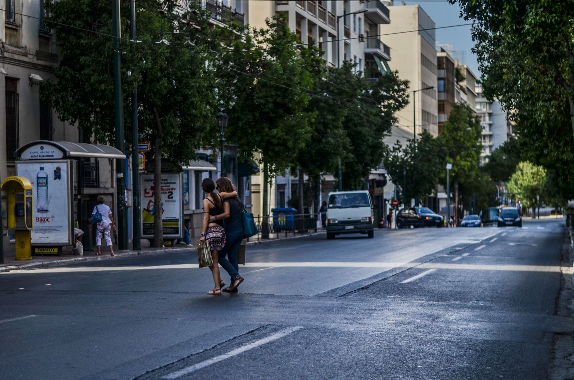 A vibrant day in Athens with people crossing a busy street lined with cars and green trees.