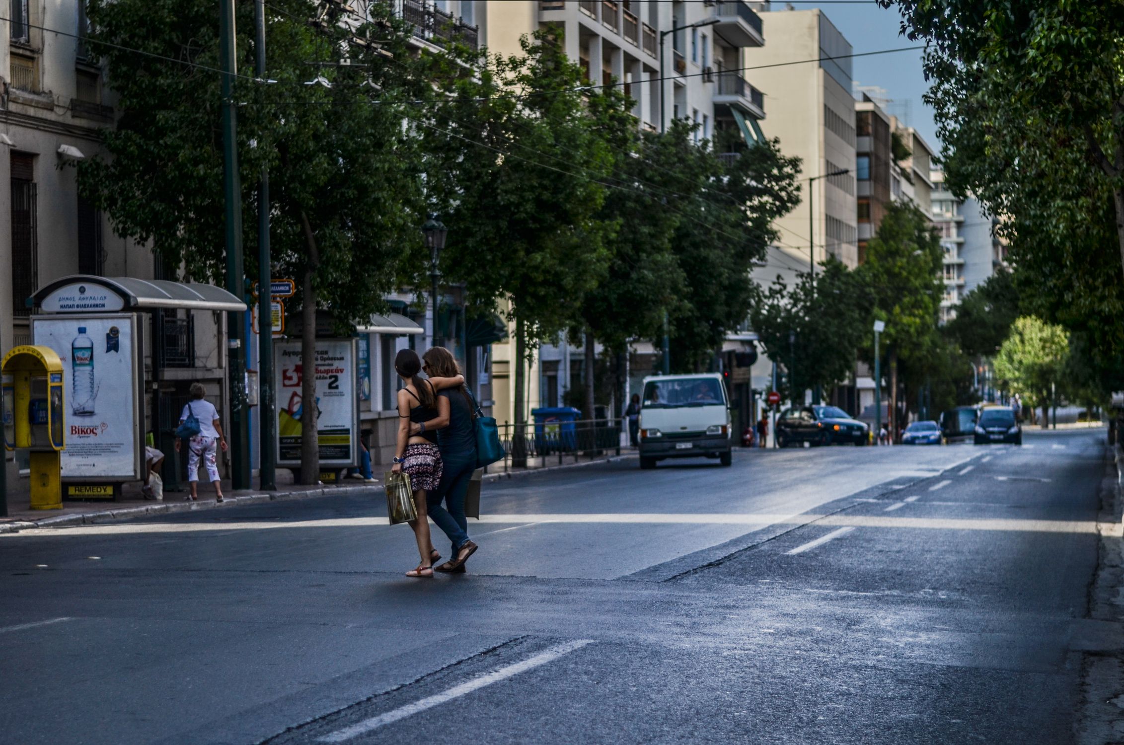 A vibrant day in Athens with people crossing a busy street lined with cars and green trees