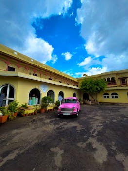 A charming scene of a vintage pink car parked in front of a historic building with a vibrant blue sky.