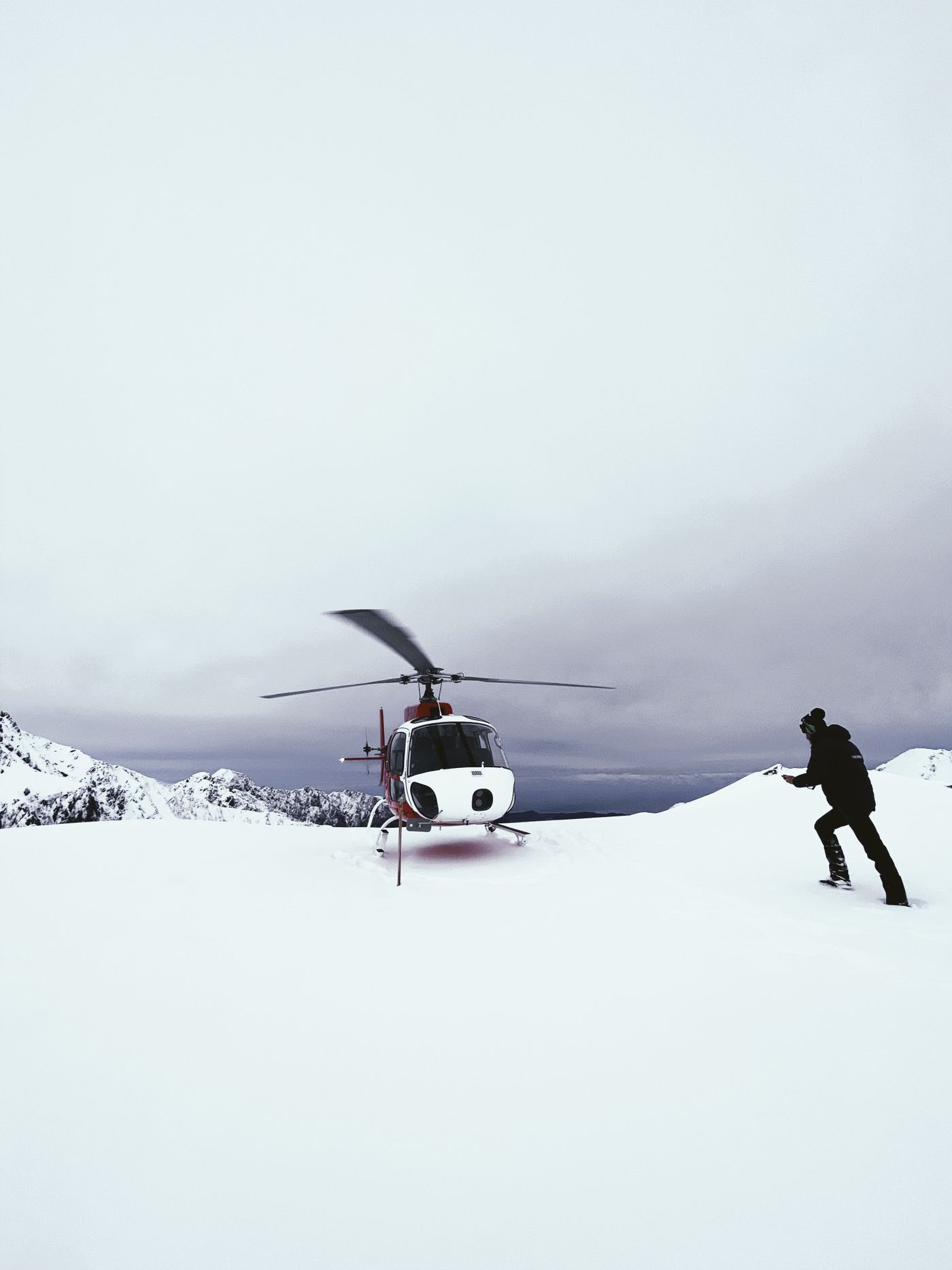 A helicopter landing on a snow-covered mountain ridge as a skier walks towards it