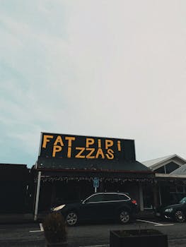 Street view of Fat Pipi Pizzas with parked cars on a cloudy day.