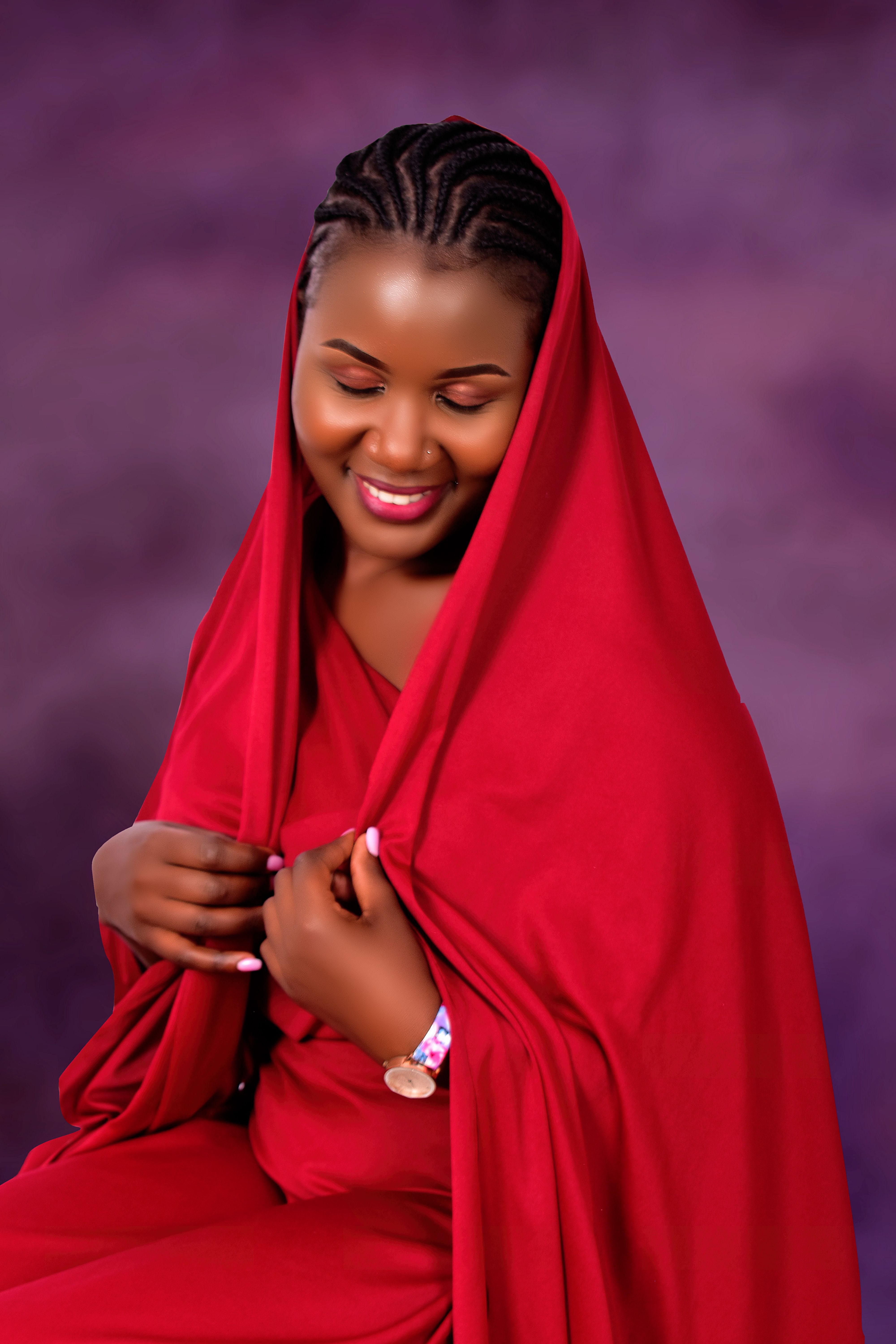 A joyful woman with braided hair wearing a red hijab, smiling gracefully in a studio shot.