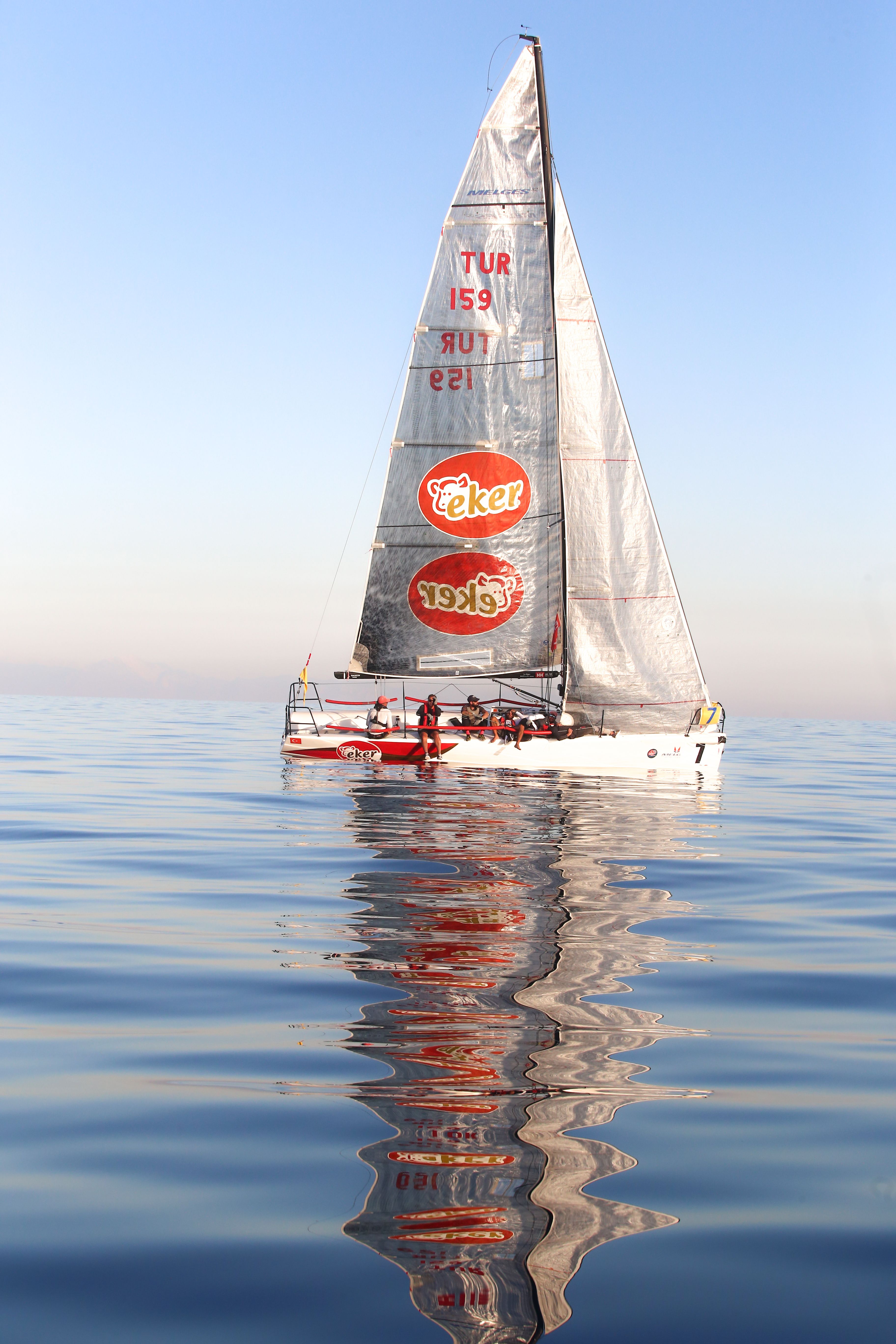 A sleek sailboat reflects on tranquil waters under a clear blue sky, perfect for sailing enthusiasts.