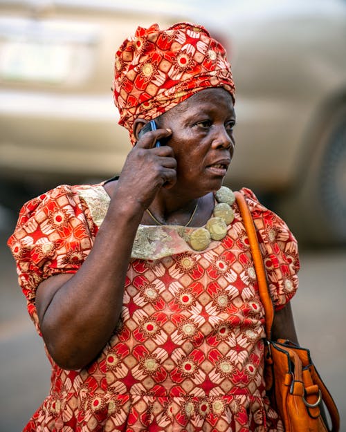 A Woman in Printed Dress Carrying an Orange Bag while Talking on the Phone