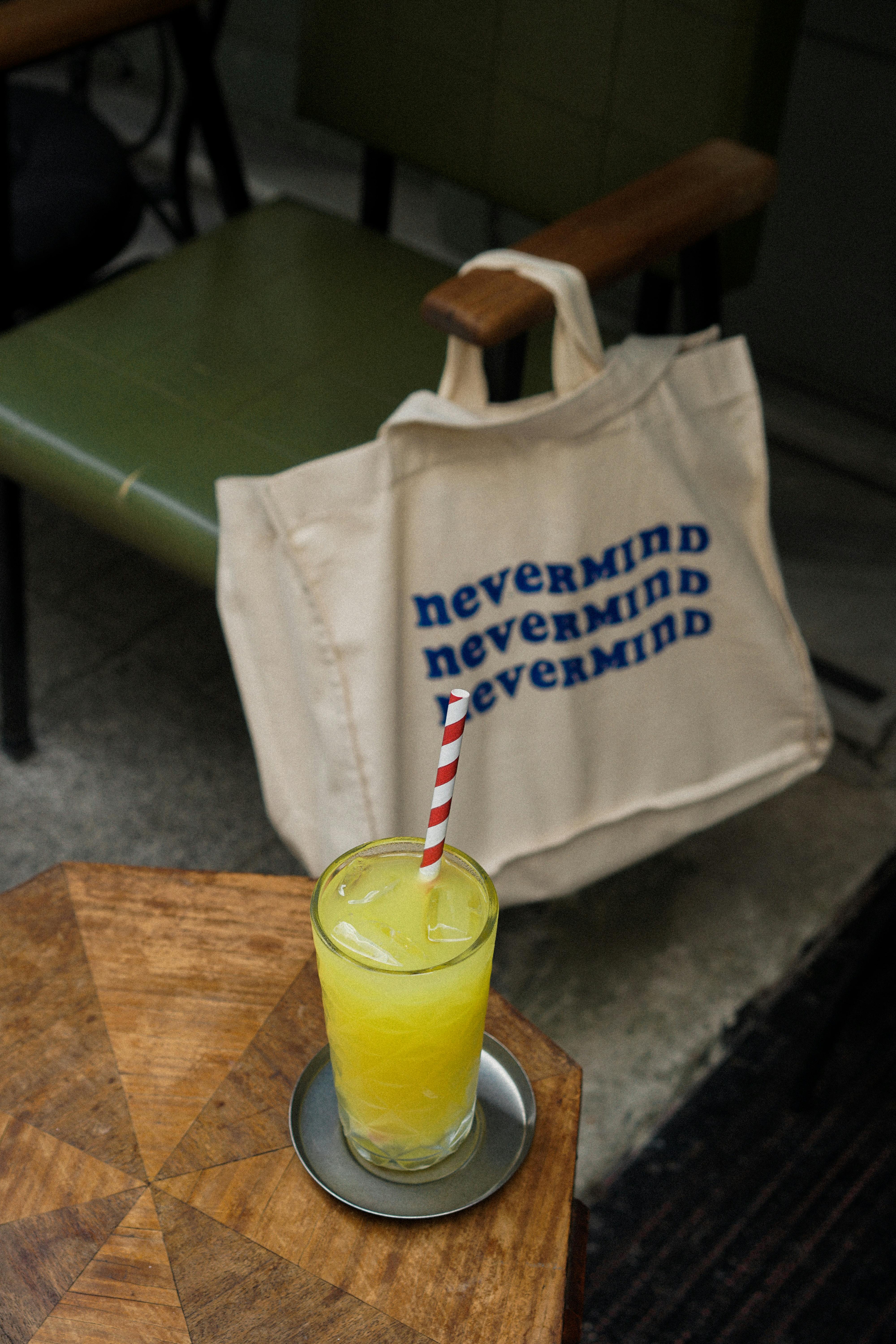 A vibrant yellow lemonade with striped straw beside a trendy tote bag on a wooden table.