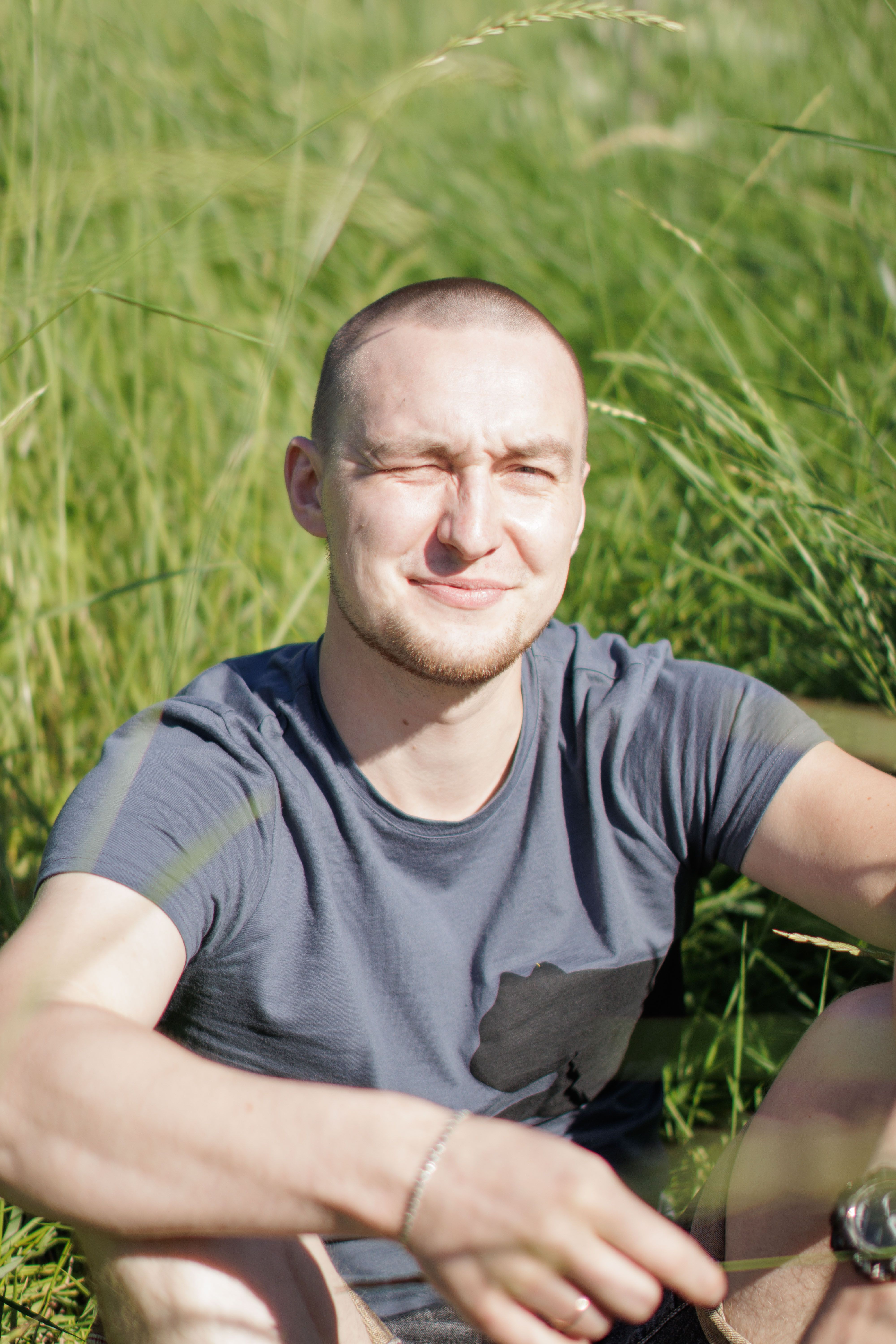 A man with a shaved head enjoys the sun in a lush green field, smiling contentedly.