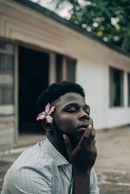 A young man with a flower in his hair, eyes closed in a serene outdoor setting.
