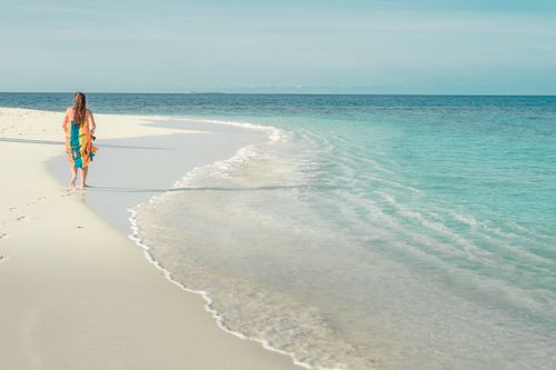 Free A woman in a colorful dress walks along a pristine white sandy beach with gentle waves. Stock Photo