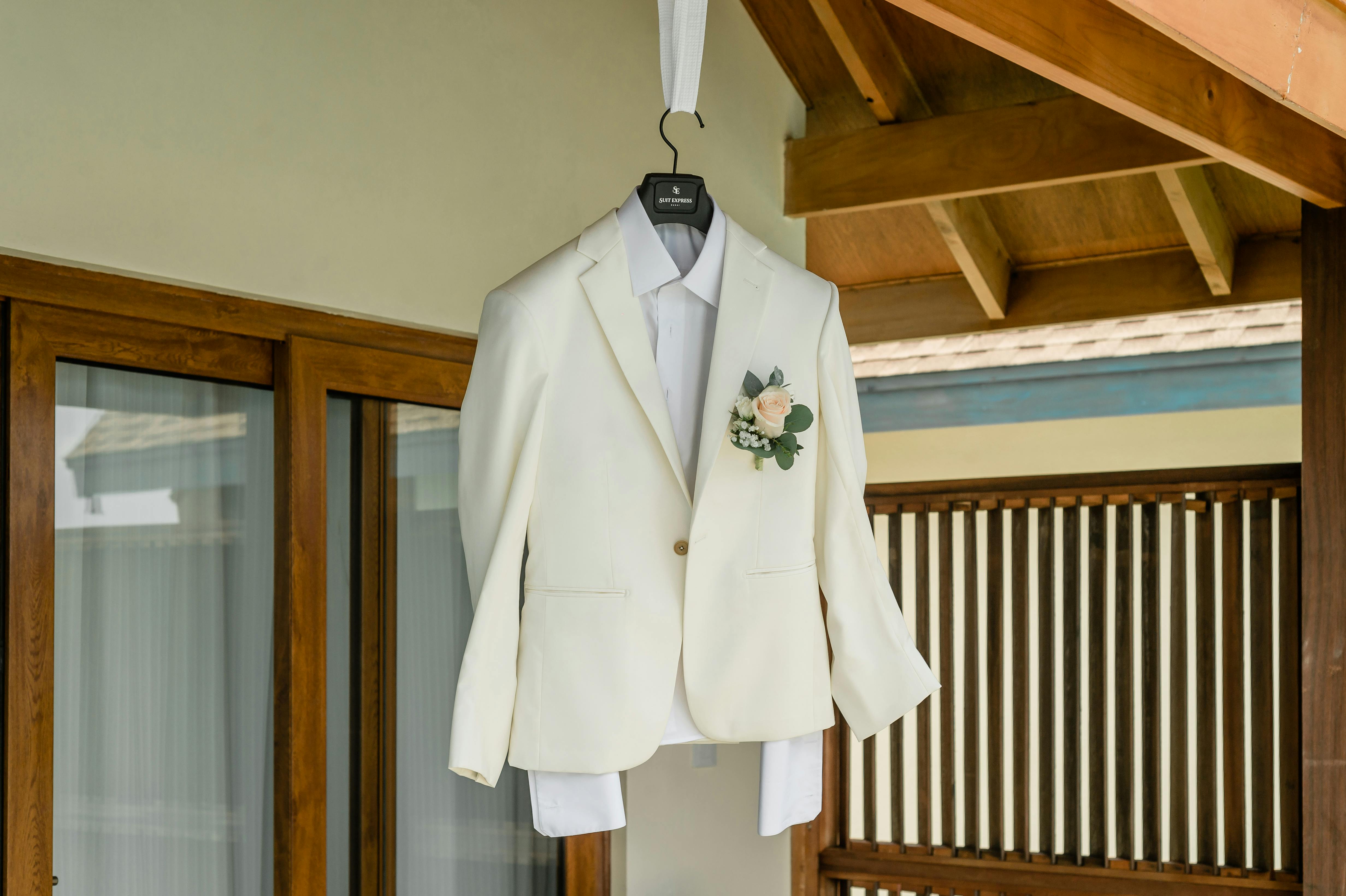 A stylish white suit jacket with a boutonniere hanging in a wooden frame setting, perfect for weddings.
