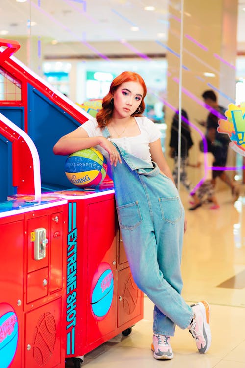 A Woman in White Shirt and Denim Jumper Leaning on Basketball Machine Arcade while Posing at the Camera