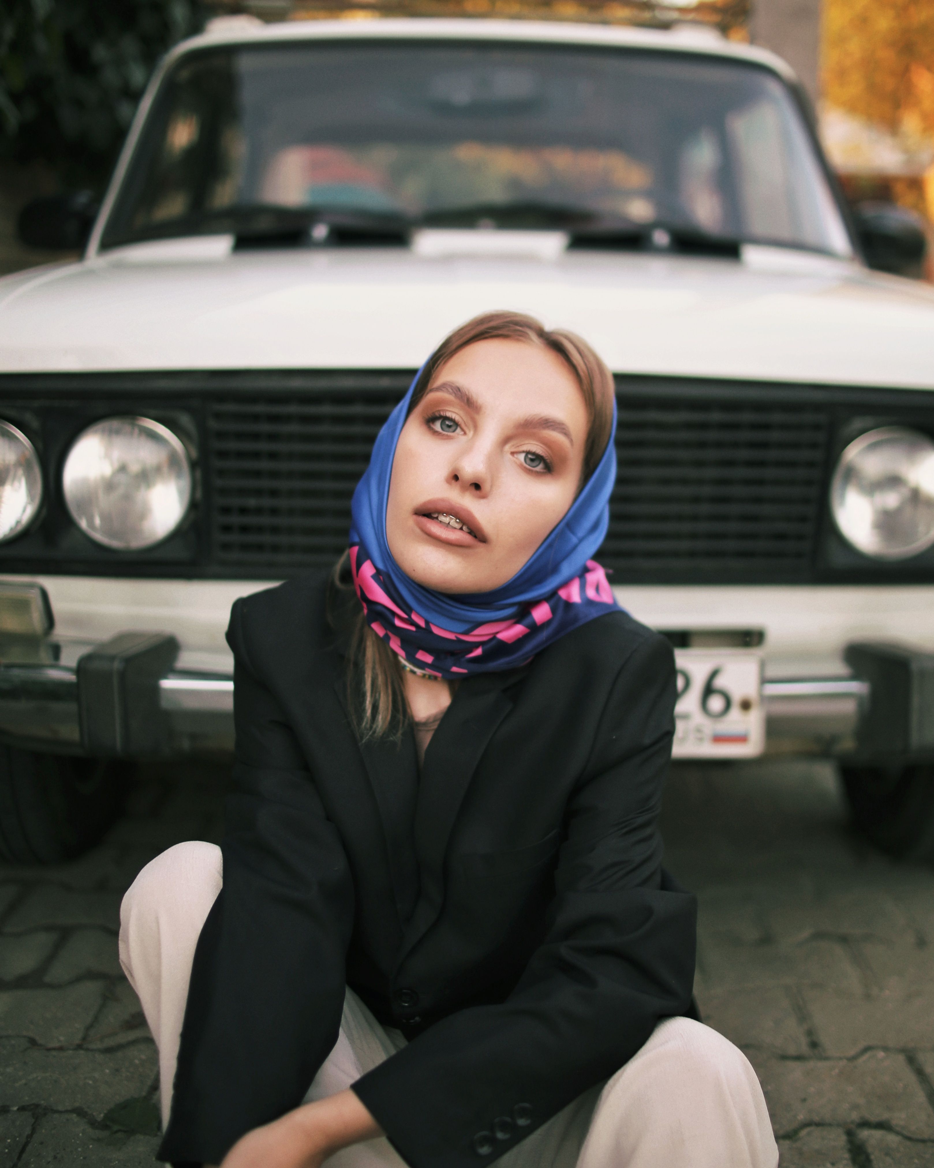 Stylish woman crouching in front of a vintage car, wearing a headscarf and blazer.