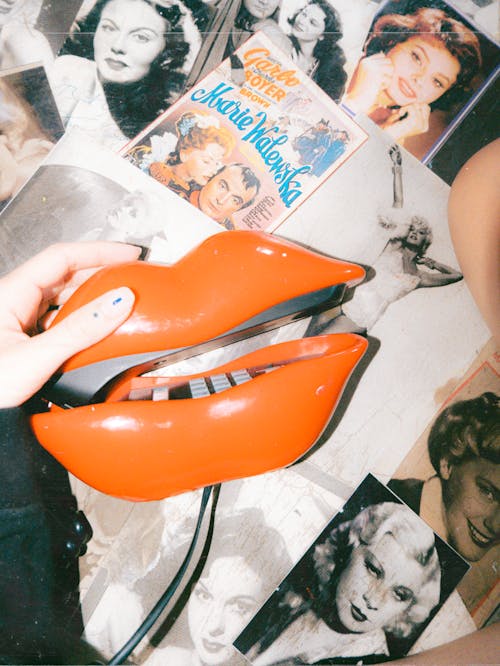 Woman Holding a Receiver of a Vintage Telephone in a Shape of Lips Lying on the Background of Scattered Photographs 