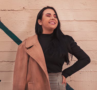 A happy woman in stylish attire smiles with confidence against a concrete wall.