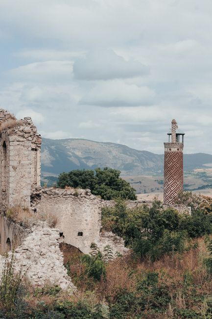 Explore ancient stone ruins and a historic tower in a mountainous landscape under a cloudy sky.
