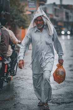 Senior man with plastic bag walking on rain-soaked street, covering head with cloth.