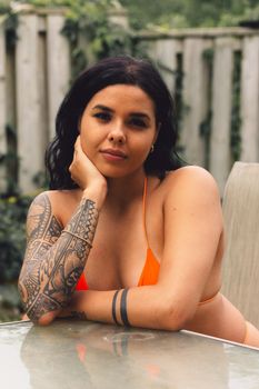 Tattooed woman in orange bikini lounging at a table outdoors. Captivating and relaxed pose.