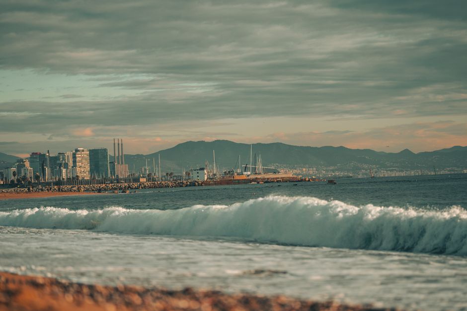 Dramatic waves crashing on a beach with a city skyline and mountains in the background at sunset.