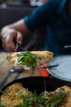 From above of crop anonymous male showing piece of tasty Italian pie with melted cheese and golden surface decorated with fresh arugula leaves while sitting at table in cafe