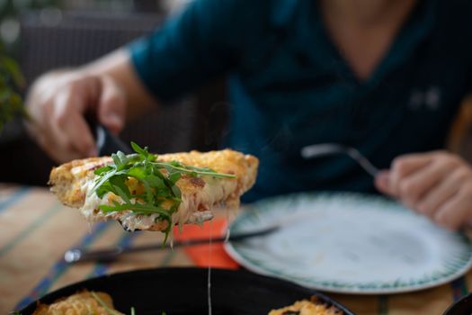 Crop unrecognizable guy serving yummy piece of pie with cheese and fresh arugula leaves while sitting with fork and plate at table in cafe