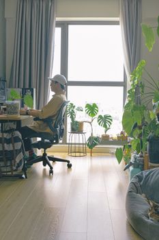 A man in a cap works on a computer in a cozy home office filled with lush indoor plants.