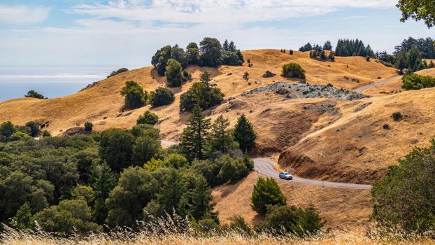 A car on a picturesque winding road amidst rolling golden hills and lush greenery.