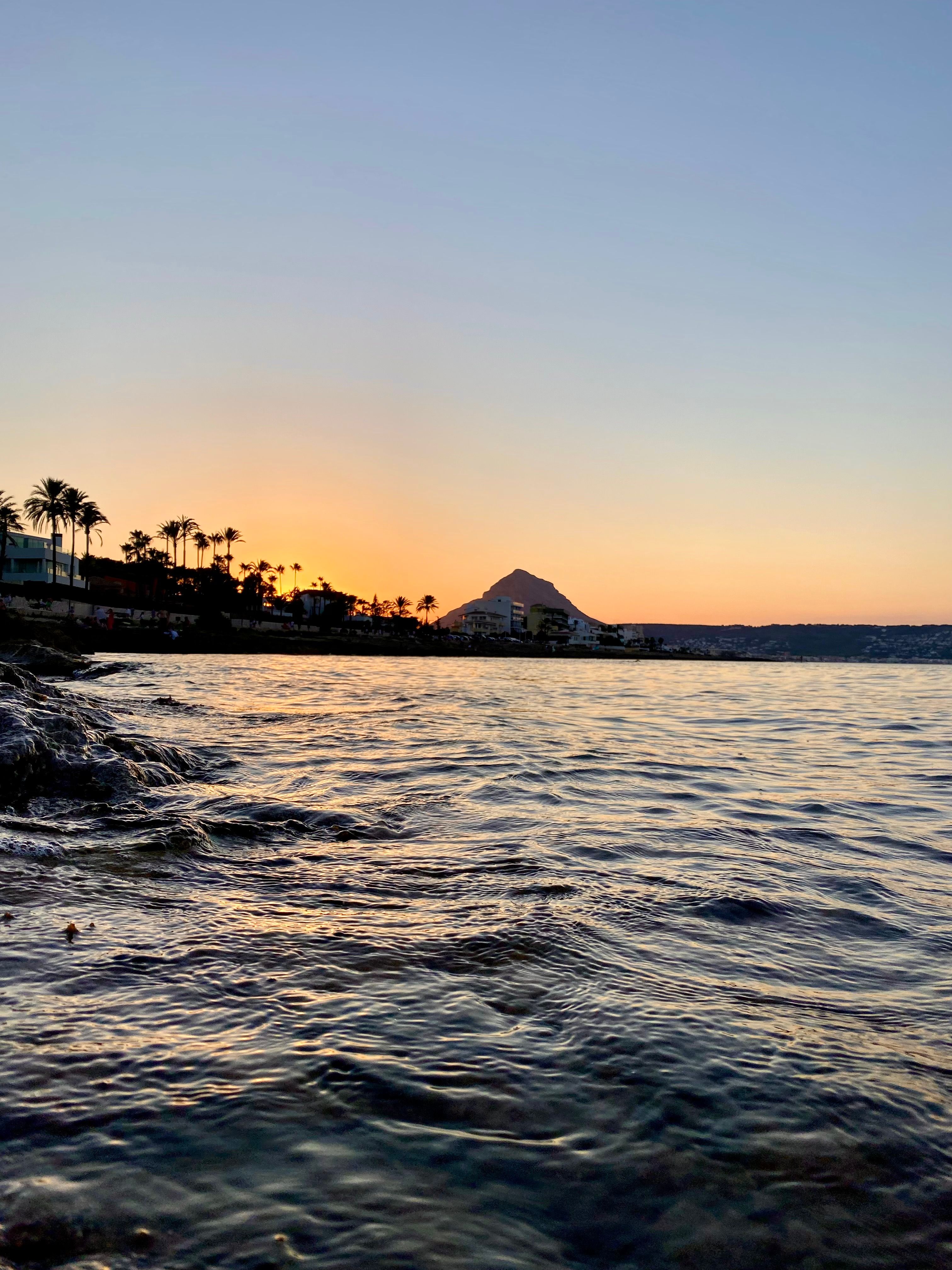Serene sunset view of the Mediterranean Sea in Xàbia, Spain with distant mountains and palms.
