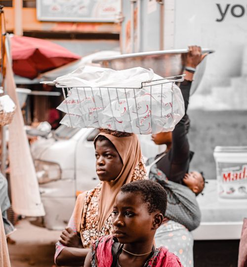 Free A woman and child in Accra market carrying goods with traditional headscarves, vibrant urban setting. Stock Photo