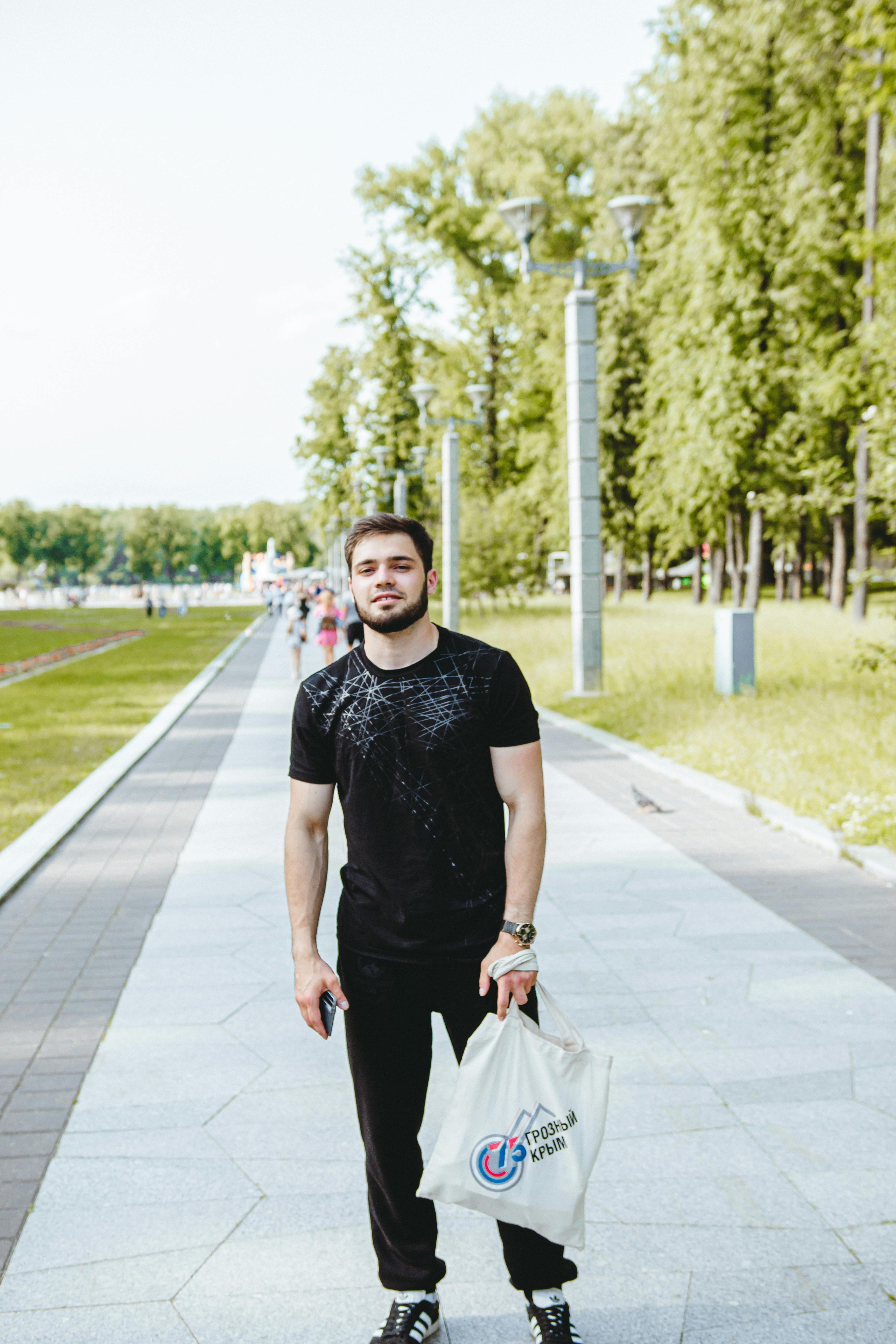 Casual young man standing on a sunny summer sidewalk holding a shopping bag.