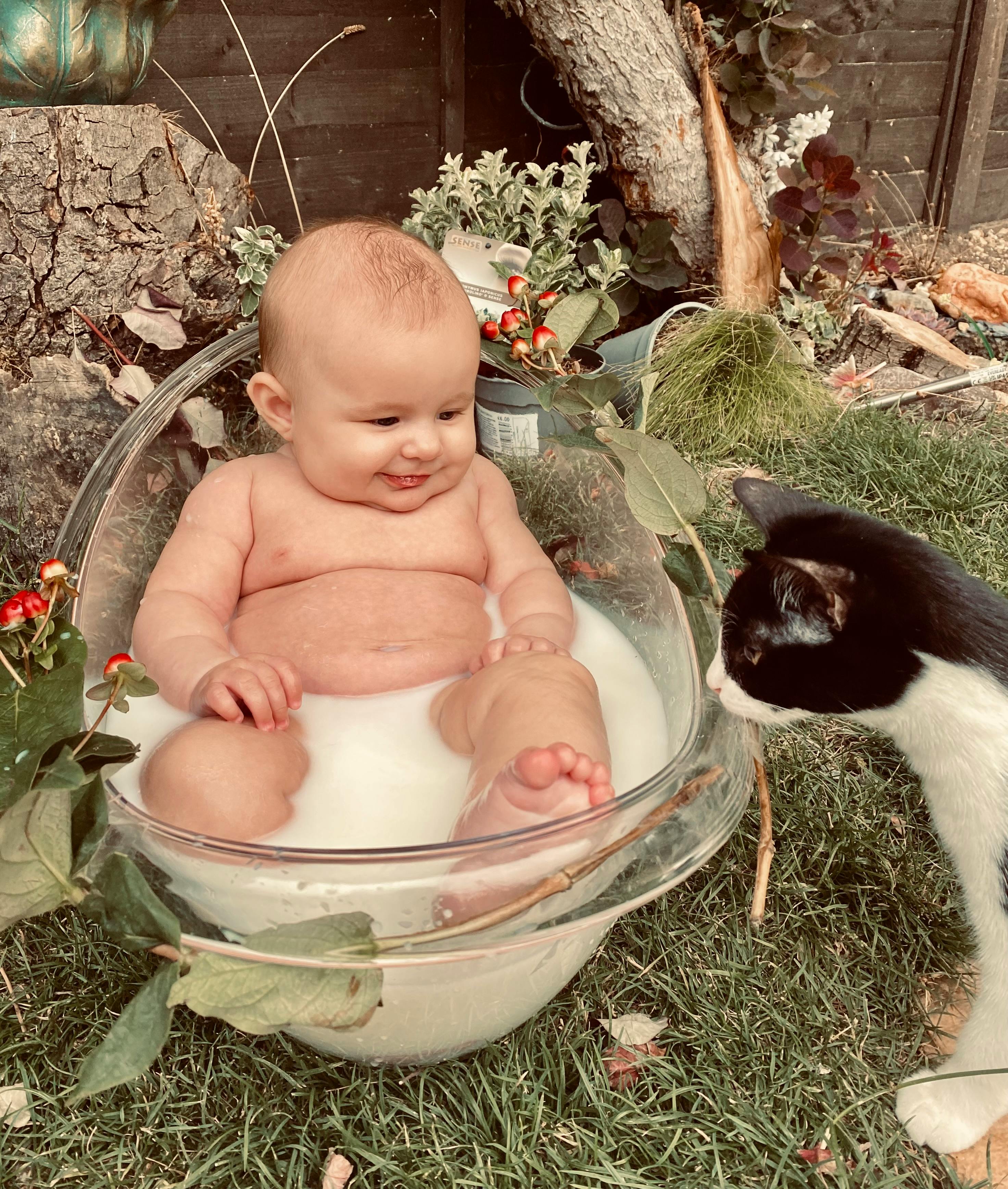 A delightful baby in a milky bath outdoors, interacting with a curious cat.