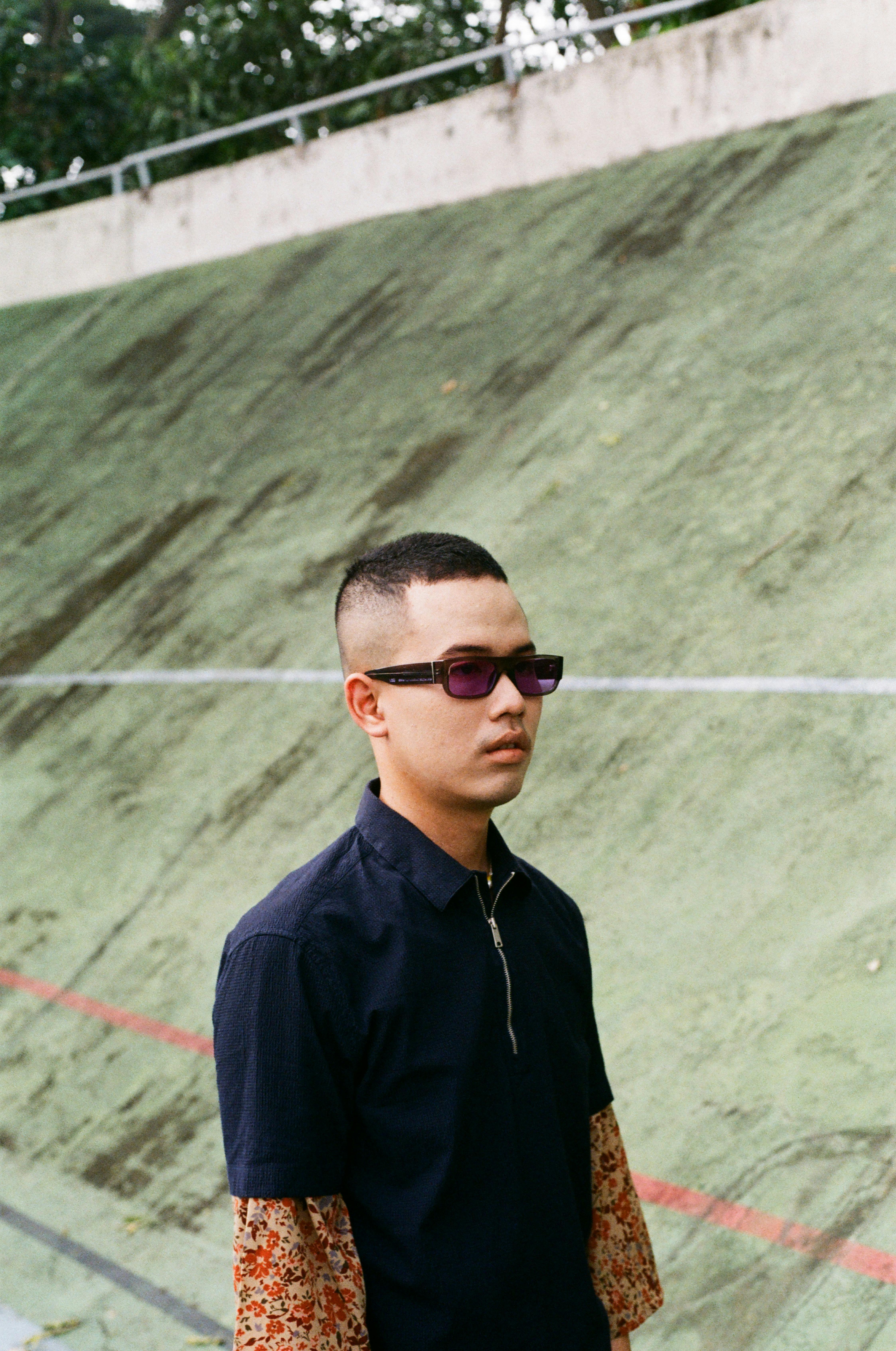 Asian man in sunglasses with serious expression standing against sloped background outdoors.