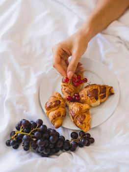A close-up of a hand picking berries from a plate with croissants, accompanied by grapes on a white linen.