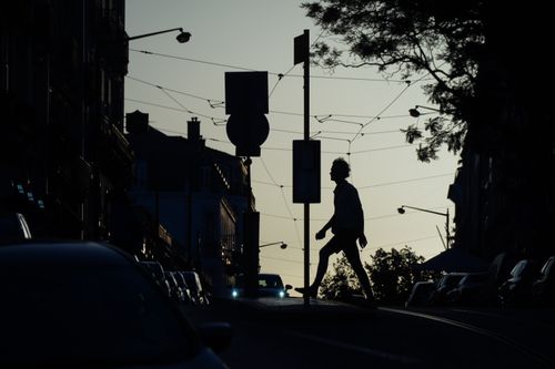 Free Silhouette of a person walking across a city street with cars and trees during sunset. Moody urban atmosphere. Stock Photo