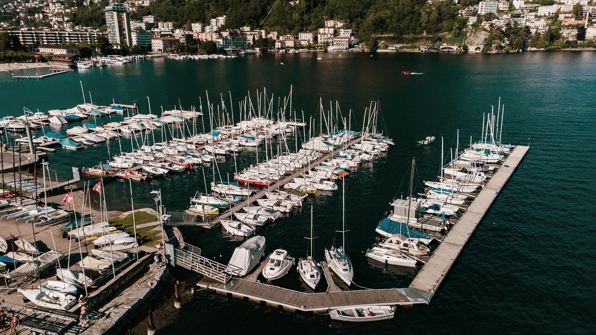 Explore an aerial view of boats docked at Lugano marina with vibrant city backdrop.