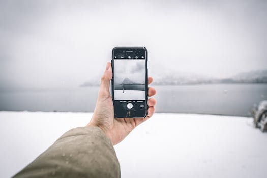 Close-up of a hand holding a smartphone capturing a serene snowy lake view.