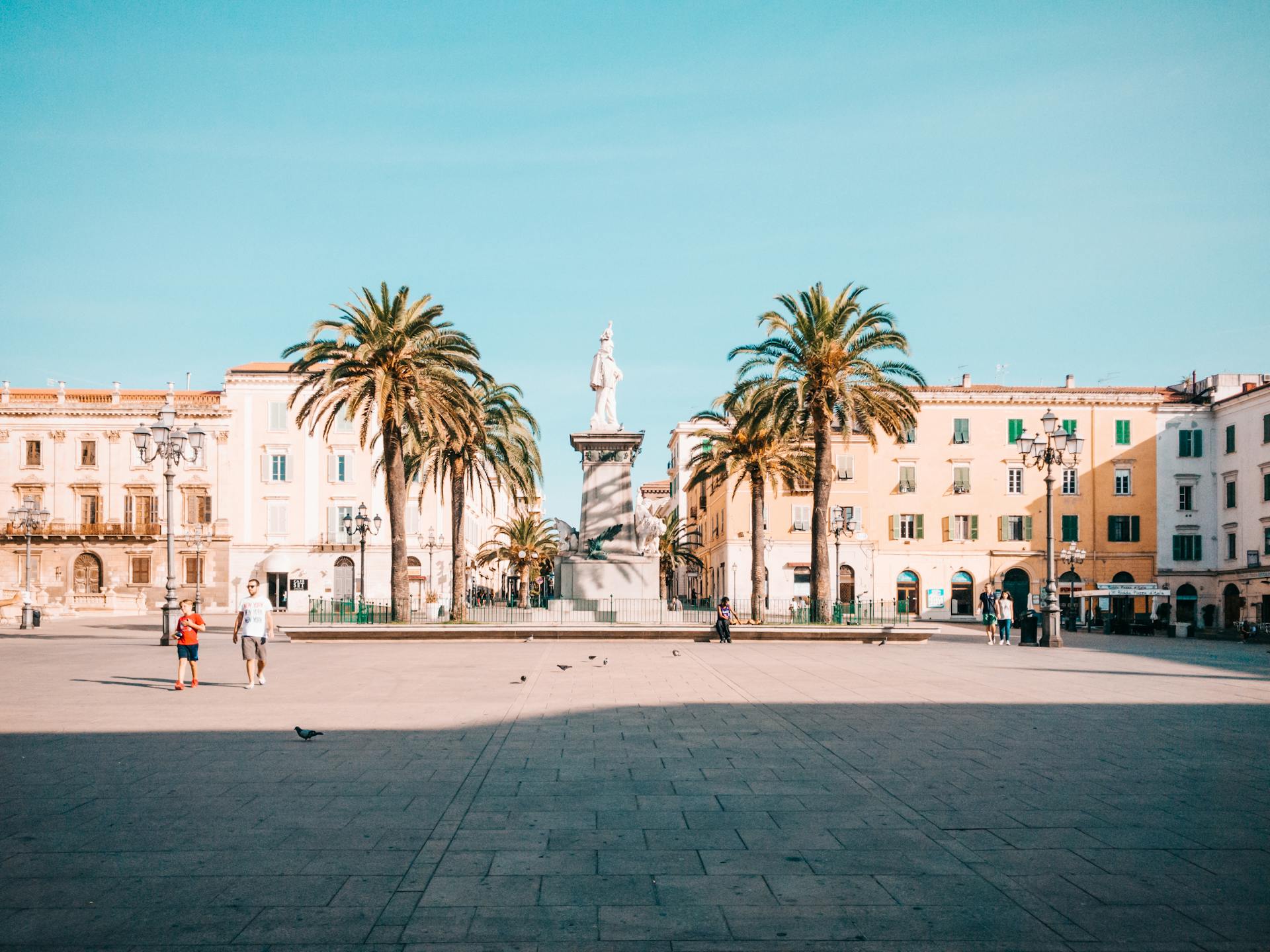 Scenic view of Piazza d'Italia in Sassari, Sardinia, showcasing palm trees, statues, and historic architecture.