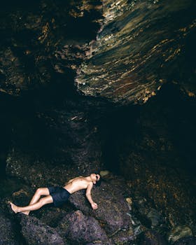 A shirtless man in shorts relaxes in a rocky cave, showcasing natural textures and shadows.