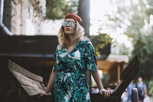 Free Young woman in a tropical print jumpsuit with sunglasses in a relaxed outdoor setting. Stock Photo