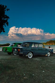 Vintage cars parked by a lake under a vibrant sunset sky, capturing a nostalgic moment outdoors.