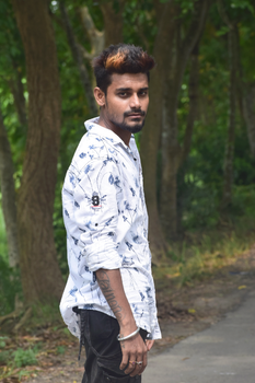 A fashionable man with floral shirt posing in Shyamnagar's lush forest area, showcasing modern style and tattoos.