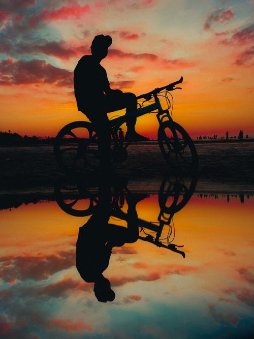Free A cyclist silhouetted against a vibrant sunset, reflected in a puddle. Stock Photo