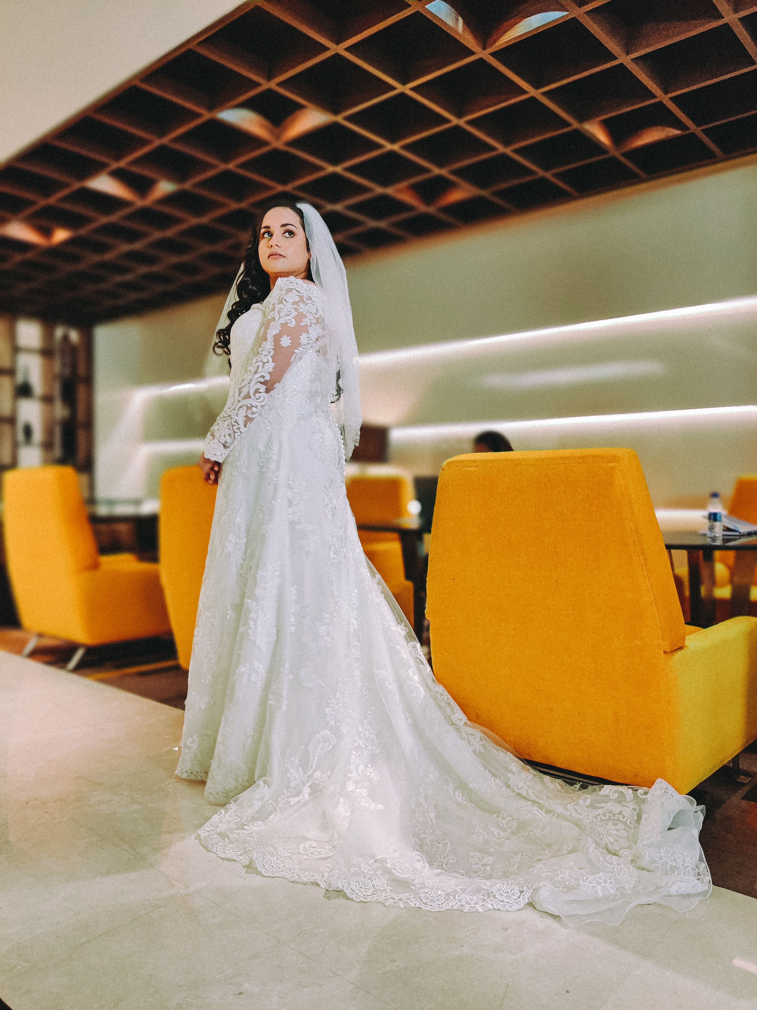 A beautiful bride in a white lace gown stands indoors with vibrant yellow chairs, exuding elegance on her wedding day.