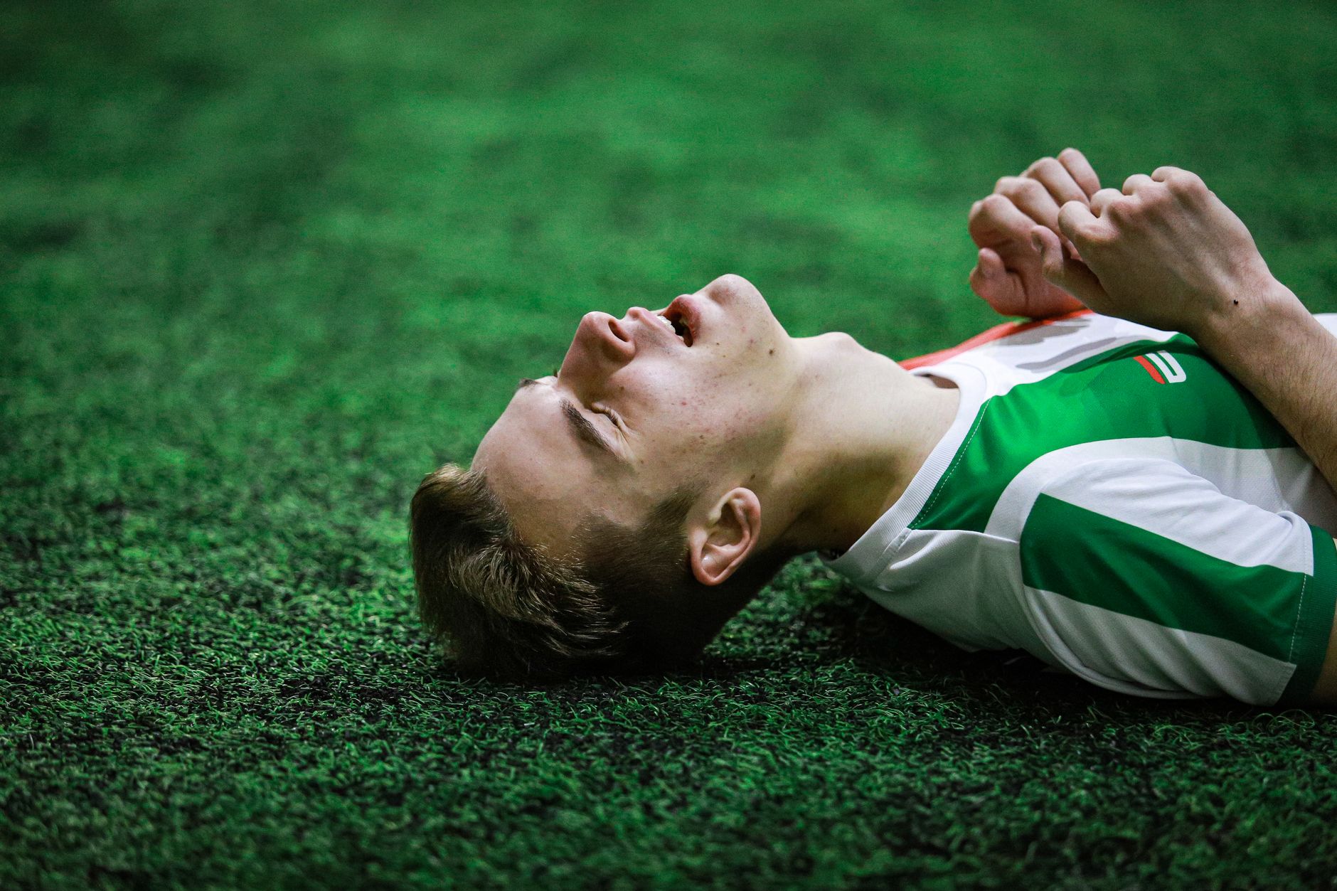 Young male soccer player in green jersey lying on grass, eyes closed, exhausted.